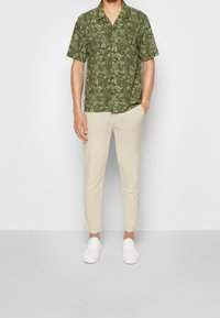 Camicia a maniche corte con bottoni di colore verde scuro con stampa a foglie, abbinata a pantaloni beige chiaro, a gamba affusolata e sneakers bianche.