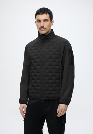 Homme aux cheveux courts et foncés et à moustache portant une veste noire matelassée avec fermeture éclair et poches, debout devant un fond clair et uni.