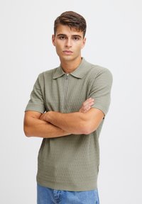 Kortermet polo skjorte i olivengrønn, strukturert strikket stoff, har krage med hel glidelås og ribbestrikket kant. Båret med blåjeans.