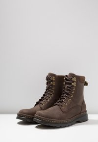 Botas de cuero marrón hasta el tobillo con cordones, punta redonda y ojales en tono dorado. Cuentan con un acabado texturizado y una suela de goma negra contrastante.