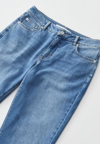 Jean en denim bleu avec poches avant, passants de ceinture et fermeture par bouton métallique posé à plat sur une surface blanche.