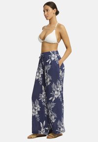 Een vrouw in een wit bikini-top draagt marineblauwe bloemen-brede broek en sandalen, poseert zijwaarts met een zelfverzekerde uitstraling.
