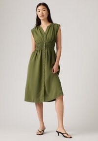 Olīvu zaļš pogājams kleita ar īsām piedurknēm, smagotu jostas vietu un potītes garumu. Gaisīga auduma ar gludu tekstūru. Melni flip-flopi.