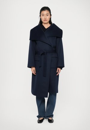 Cappotto avvolgente blu navy con ampia colletto, chiusura frontale con cravatta e due tasche laterali. Abbinato a jeans blu e scarpe nere. Texture liscia.