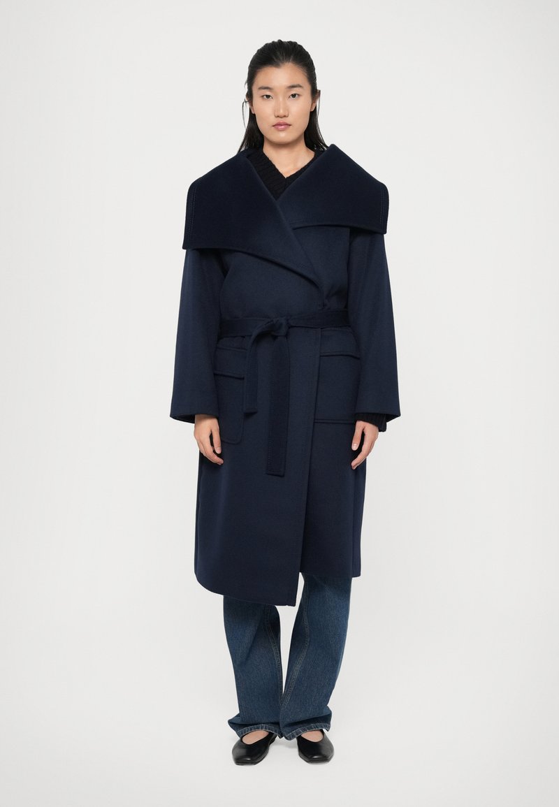 Manteau en bleu marine avec un large col, fermeture par lien à l'avant et deux poches latérales. Associé à un jean bleu et des chaussures noires. Texture lisse.