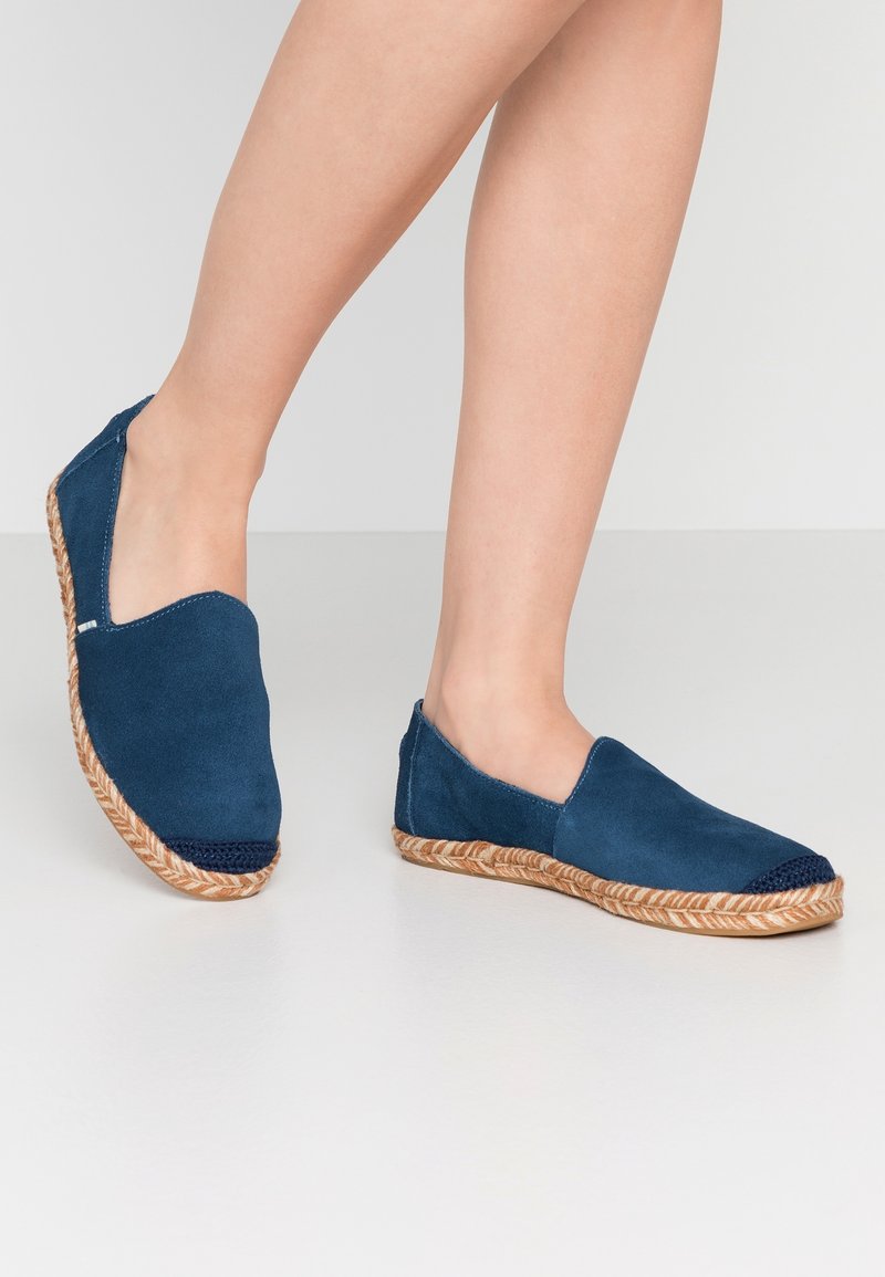 Jambes portant des espadrilles bleues slip-on avec des semelles en jute tressé et des bouts d’orteils sombres, debout sur une surface blanche unie.