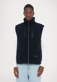 Gilet en polaire marine avec col montant, zip frontal et deux poches poitrine zippées. Il présente un tissu texturé et un petit logo sur l'ourlet.