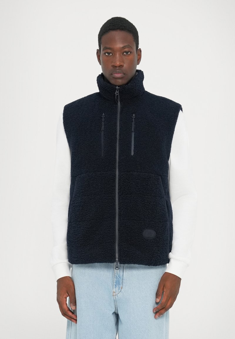 Gilet en polaire marine avec col montant, zip frontal et deux poches poitrine zippées. Il présente un tissu texturé et un petit logo sur l'ourlet.