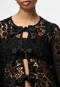 Kvinne som har på seg en svart blomstermønstret blonderbluse med tre svarte satengsløyfer knyttet langs foran.