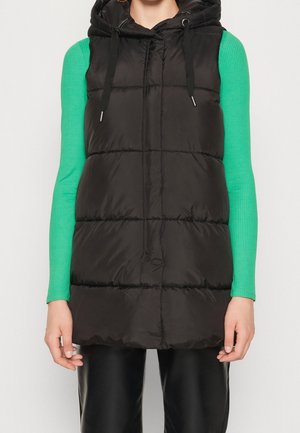 Persona que lleva un chaleco negro con capucha sobre una camisa verde brillante de manga larga y pantalones negros, mostrado desde los hombros hasta la mitad del muslo.