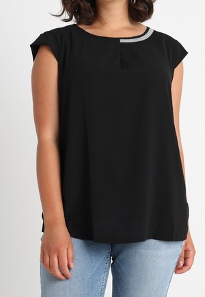 Kobieta nosząca czarną koszulkę z krótkim rękawem z małym szarym detalem przy dekolcie, w połączeniu z jasnoniebieskimi jeansami.