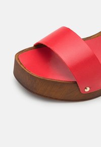 Sandale mules en cuir rouge avec large bride et semelle plateforme en bois marron sur fond blanc.