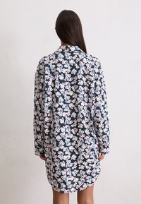 Mujer con cabello largo y oscuro, vistiendo un vestido floral de manga larga con flores rosas, blancas y azules, de pie contra una pared blanca, vista desde atrás.