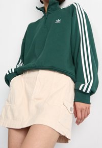 Groene fleece pullover met een halve zip-nek, met witte Adidas merknaam en drie witte strepen op de mouwen. Gecombineerd met een crèmekleurige rok.