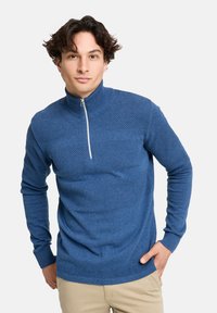 Blaues Strickpullover mit hohem Kragen und halbem Reißverschluss, ausgestattet mit strukturierten Mustern und gerippten Bündchen, getragen über hellen Hosen.