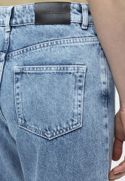 Φωτεινά γαλάζια τζιν με πίσω τσέπη και μαύρο δερμάτινο έμβλημα με την ένδειξη IVY OAK στη ζώνη, φορεμένα από άτομο με το χέρι να ακουμπά δίπλα στο ισχίο.