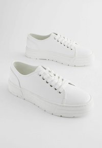 Adidași casual albi cu un meshed superior sintetic neted, vârf rotund, talpă plată și gauri din metal pentru șireturi. Design și textură minimaliste.