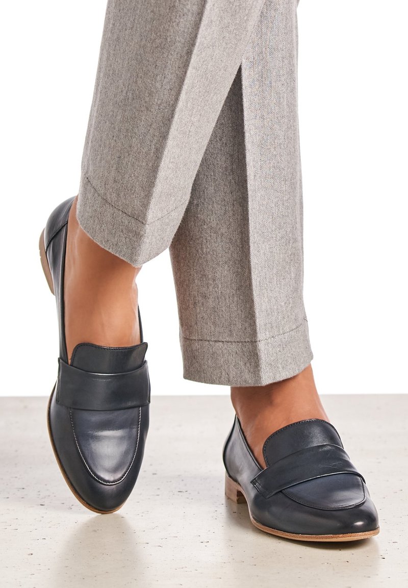 Mocassins en cuir bleu marine à finition lisse, avec une large bande supérieure et un petit talon, associés à un pantalon gris clair et ajusté.