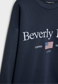 Sweat-shirt bleu marine avec le mot « Beverly » brodé en blanc, un petit drapeau américain et les inscriptions « United » et « 1999 » sous le logo principal.