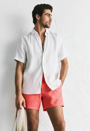 White short-sleeve linen shirt, paired with coral-red textured shorts featuring vertical stripes. Model holds a beige tote bag.