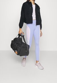 Svart huva jacka, ljuslila och blå mönstrade leggings, rosa sneakers och en svart duffelväska med en orange logotyp.