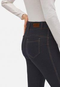 Mujer con jeans ajustados de color azul oscuro con costuras naranjas visibles y una etiqueta de cuero marrón, combinados con una blusa de manga larga blanca.