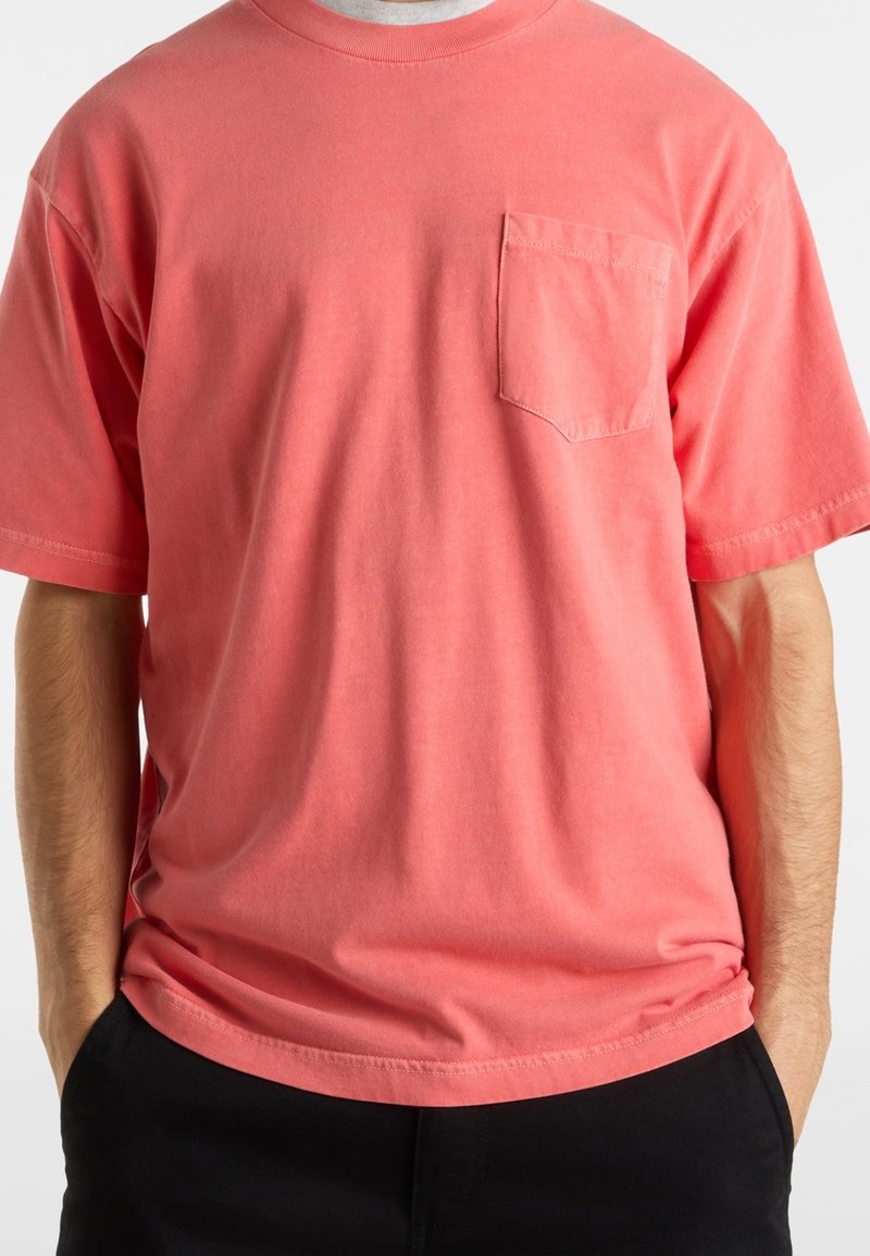 Homme portant un t-shirt corail à manches courtes avec une poche poitrine gauche et un pantalon noir, mains dans les poches, sur fond blanc.