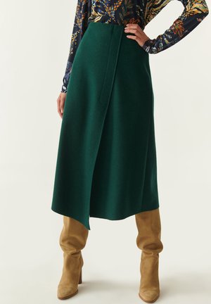 Vrouw in een donkergroene asymmetrische midi-rok, met een geprinte lange mouw top en tan suède kniehoge laarzen, staand met hand op heup.