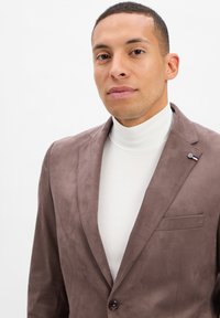 Brauner Wildleder-Blazer mit Reverskragen, einem einreihigen Knopfverschluss und einer Brusttasche. Tragen über einem weißen Rollkragenpullover. Glatte Textur, taillierter Schnitt.