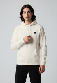 White hoodie with adjustable drawstring, front pocket, ribbed cuffs, and small black logo patch on the chest. Soft, smooth texture.