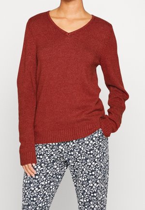 Person wearing a rust red V-neck sweater with long sleeves and black-and-white floral patterned pants, standing with one hand in pocket.