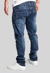 Denim jeans in dark blue, slim fit, featuring multiple pockets and subtle fading. Paired with white sneakers.