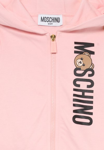 Haut zippé rose avec une fermeture éclair dorée, arborant un graphique d'ours brun et l'inscription en noir "MOSCHINO". Tissu doux avec une texture lisse.