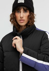 Schwarze Pufferjacke mit hohem Kragen, mit lila Akzenten und weißen Streifen an den Ärmeln; Modell trägt eine schwarze Strickmütze mit Label.