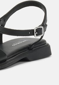 Sandalo in pelle nera con cinturino alla caviglia regolabile, fibbia quadrata e suola piatta. Caratterizzato da una superficie liscia e un design minimalista.