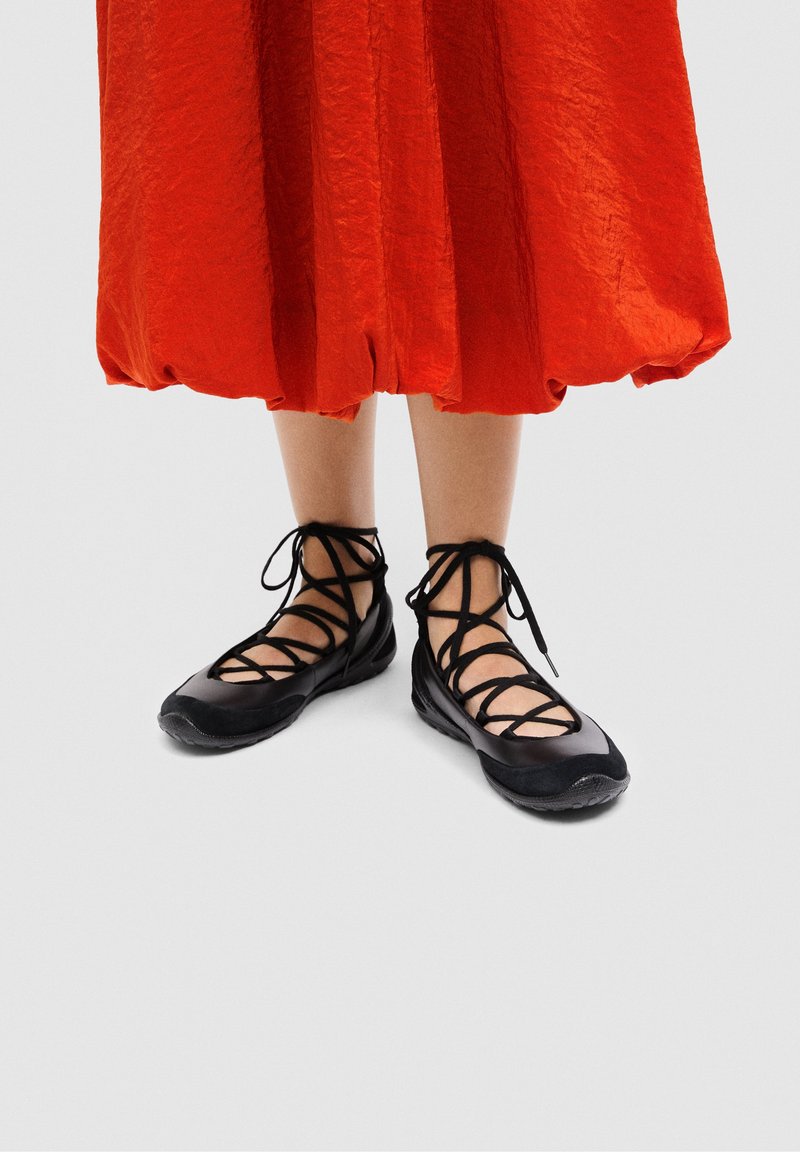 Jambes portant des ballerines noires à lacets associées à une jupe midi orange vif avec un ourlet froncé sur un fond uni.