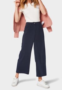 Kobieta w białych sportowych butach, granatowych szerokich spodniach, białej koszulce, trzymająca różową kurtkę zarzuconą na ramiona.