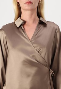 Camisa de seda castanha com gola, apresentando mangas longas e um laço lateral, exibindo uma textura suave e brilhante.