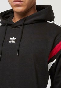 Čierna mikina s kapucňou a viazacou šnúrkou, ktorá má biele a červené pruhy na pleciach a biele logo Adidas na hrudi. Hladká textura.