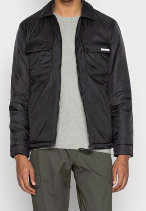 Homme portant une veste noire zippée sur une chemise grise et un pantalon cargo vert olive, debout avec les bras détendus le long du corps.