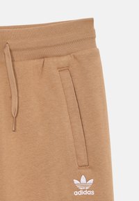 Briņas sporta bikses, kas izgatavotas no mīksta auduma, ar sānu kabatām, regulējamu jostu un baltu Adidas logotipu uz apakšējās kājas.