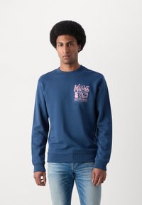 Felpa blu navy in misto cotone, con una grafica rosa sul petto sinistro e polsini e orlo a coste. Abbinata a jeans azzurri.
