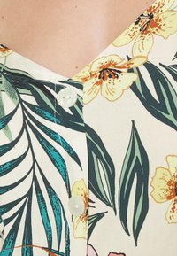 Blusa floreale con scollatura a V, tessuto leggero con foglie verdi e fiori gialli, accentuata da un bottone tondo al collo.