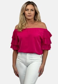 Jasnoróżowa bluzka typu off-the-shoulder z warstwowymi falbaniastymi rękawami. Gładki materiał, w zestawieniu z dopasowanymi białymi spodniami. Prosty dekolt i zwiewny krój.