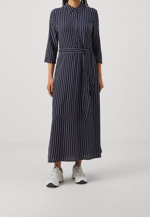 Woman wearing navy blue long-sleeve, button-up, striped dress with a waist tie and white athletic shoes, standing against a plain background.
