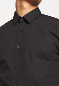 Primer plano de una persona que lleva una camisa negra de botones con un cuello puntiagudo, contra un fondo claro y liso.