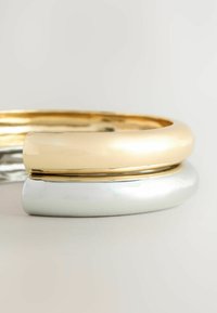 Metal armbånd i guld- og sølvtoner, med glatte, rundede former, en poleret finish og en subtil, vandret rille detaljer.