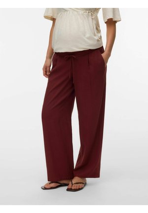 Ženska, oblečena v bordo hlače širokih nogavic z vrvico v pasu, bež top s kratkimi rokavi in črne natikače.