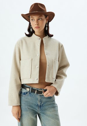 Geknipte beige fleecejack met drukknoopsluiting, twee voorzakken, gedragen over een bruine top en blauwe jeans, geaccessoireerd met een bruine hoed en oorbellen.