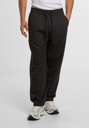 Black tapered sweatpants with a ribbed elastic waistband and drawstring, featuring side pockets and smooth texture. Worn with silver athletic shoes.